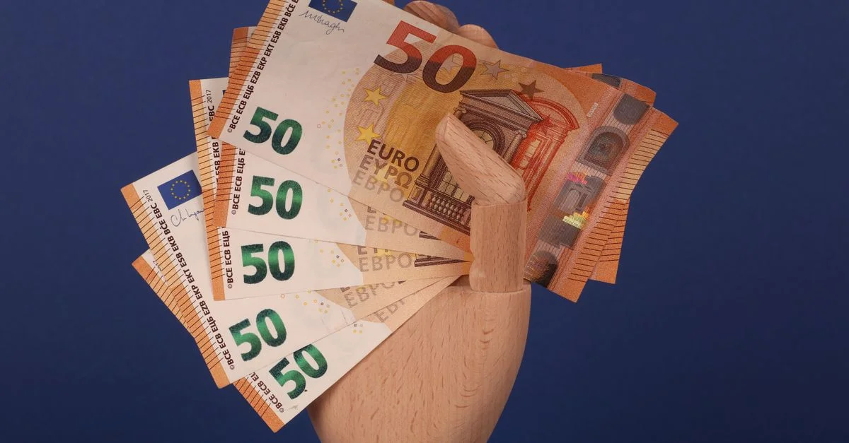 Une main en bois de mannequin tenant des billets de cinquante euros sur un fond bleu.