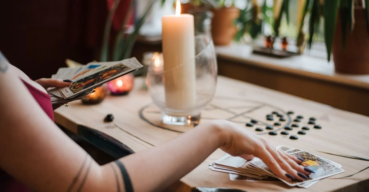 La main d'une femme arrange des cartes de tarot sur une table ornée d'une bougie et d'objets mystiques, à l'intérieur.