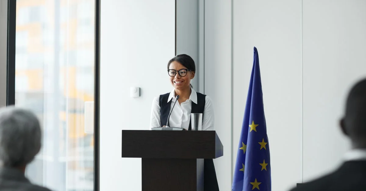 Une femme donnant un discours à un podium avec un drapeau de l'UE dans un cadre de conférence.